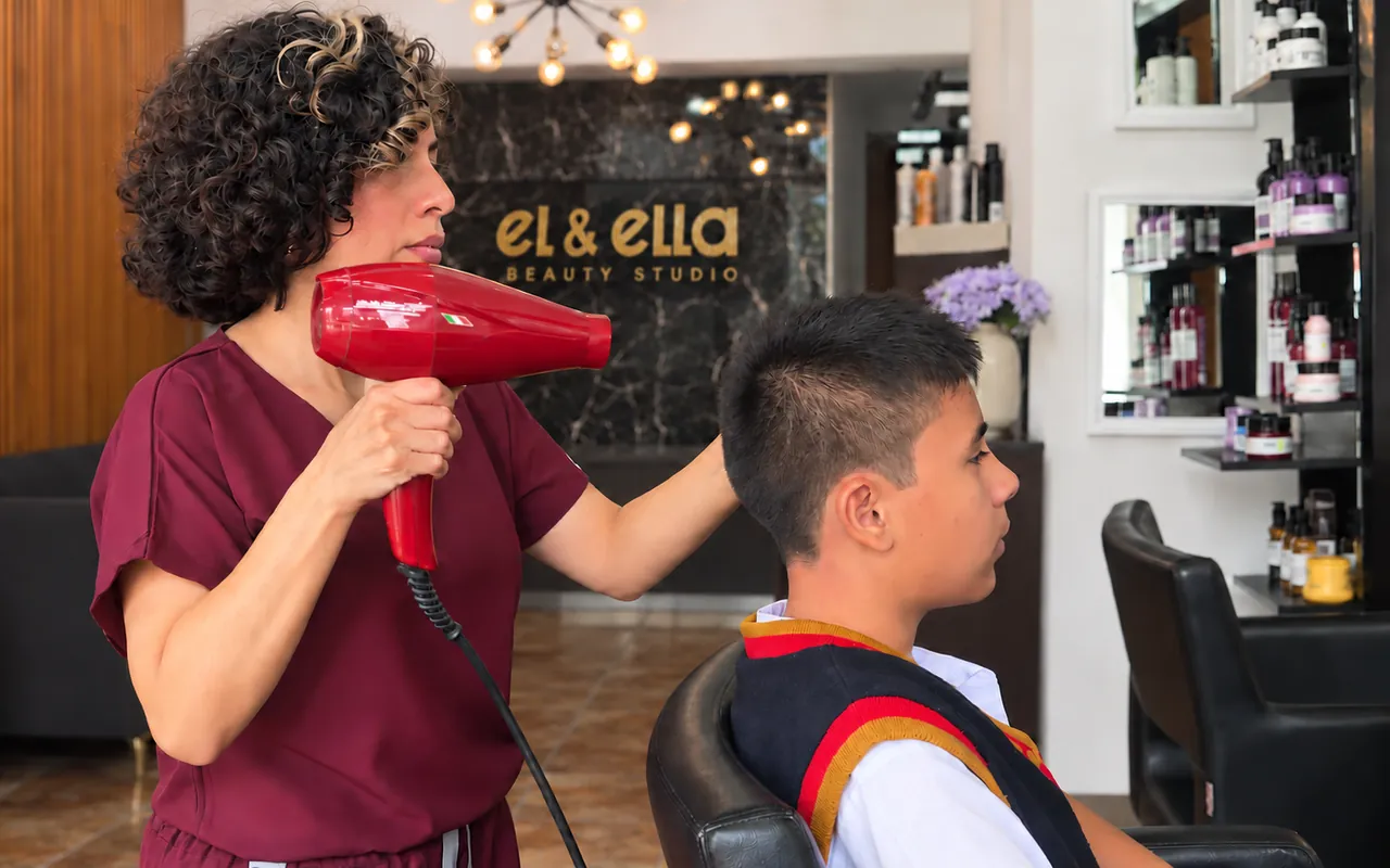Estilista del salón secando y peinando a un cliente recién cortado, con el logo dorado al fondo