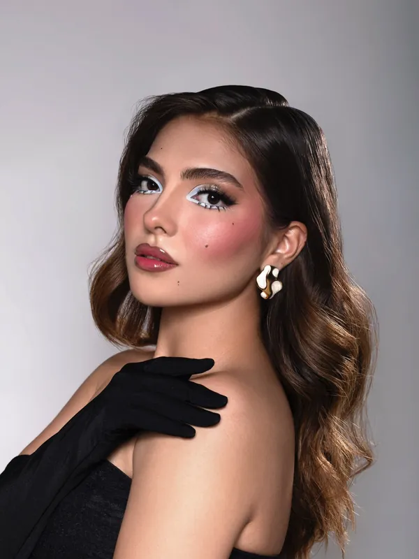 Maquillaje con delineado gráfico blanco, rubor rosado, labios brillantes y ondas suaves, de perfil con arete escultural